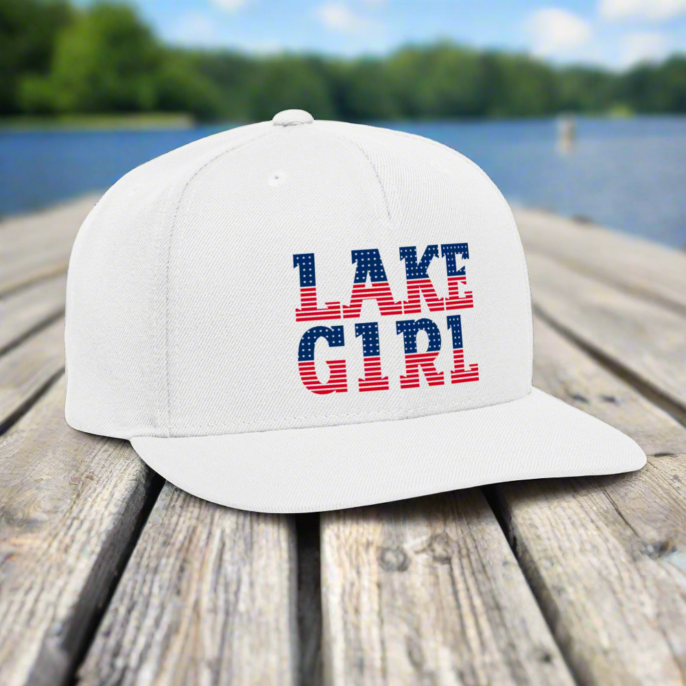 White cap with 'LAKE GIRL' text on a wooden dock by a lake